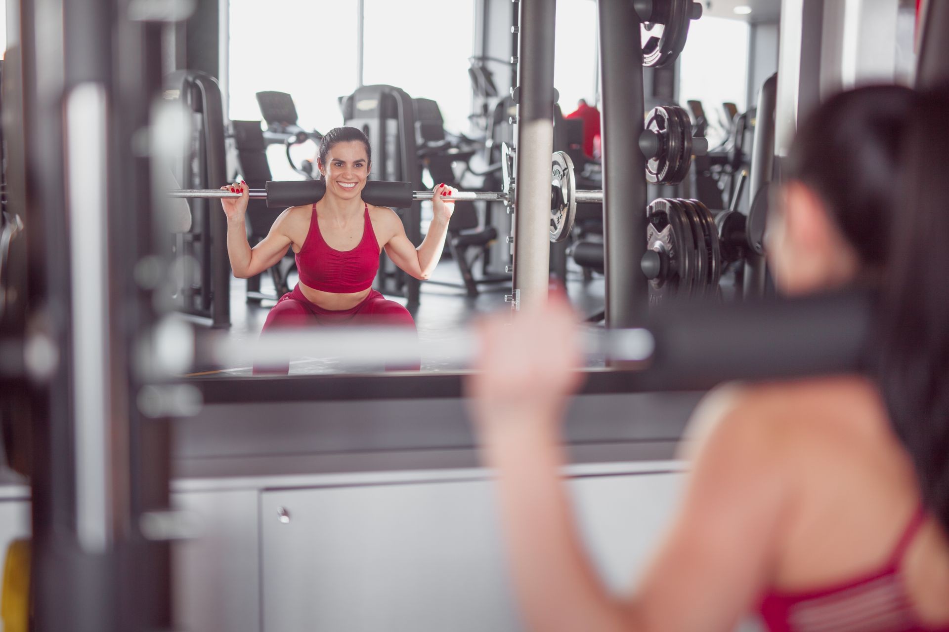 Happy middle-aged woman in gym doing squats in machine against mirror and smiling